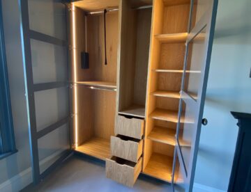 Illuminated fitted wardrobe interior with oak shelving and painted framed glass doors